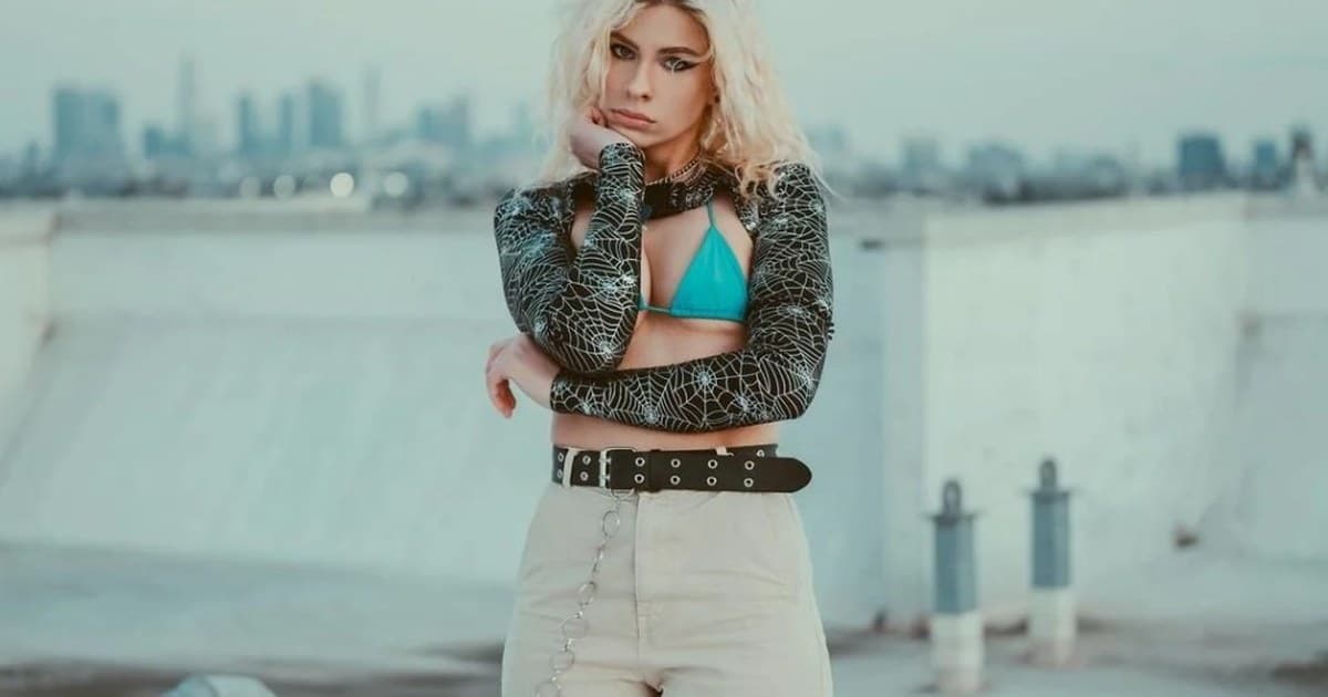 Andie Case on a rooftop with a city skyline behind her, blonde wavy hair, black spiderweb shrug, and a direct, thoughtful expression.