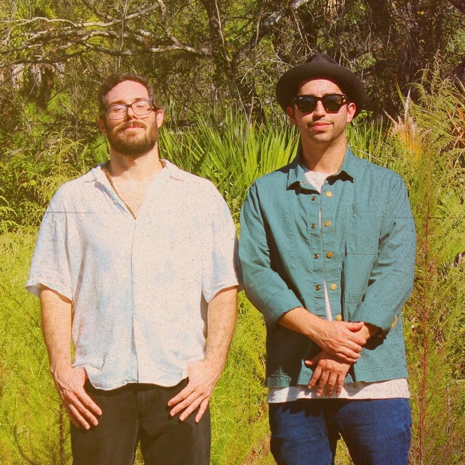 Banyan’s two members standing in sunlit Florida scrub, one in glasses and one in a black hat and sunglasses.