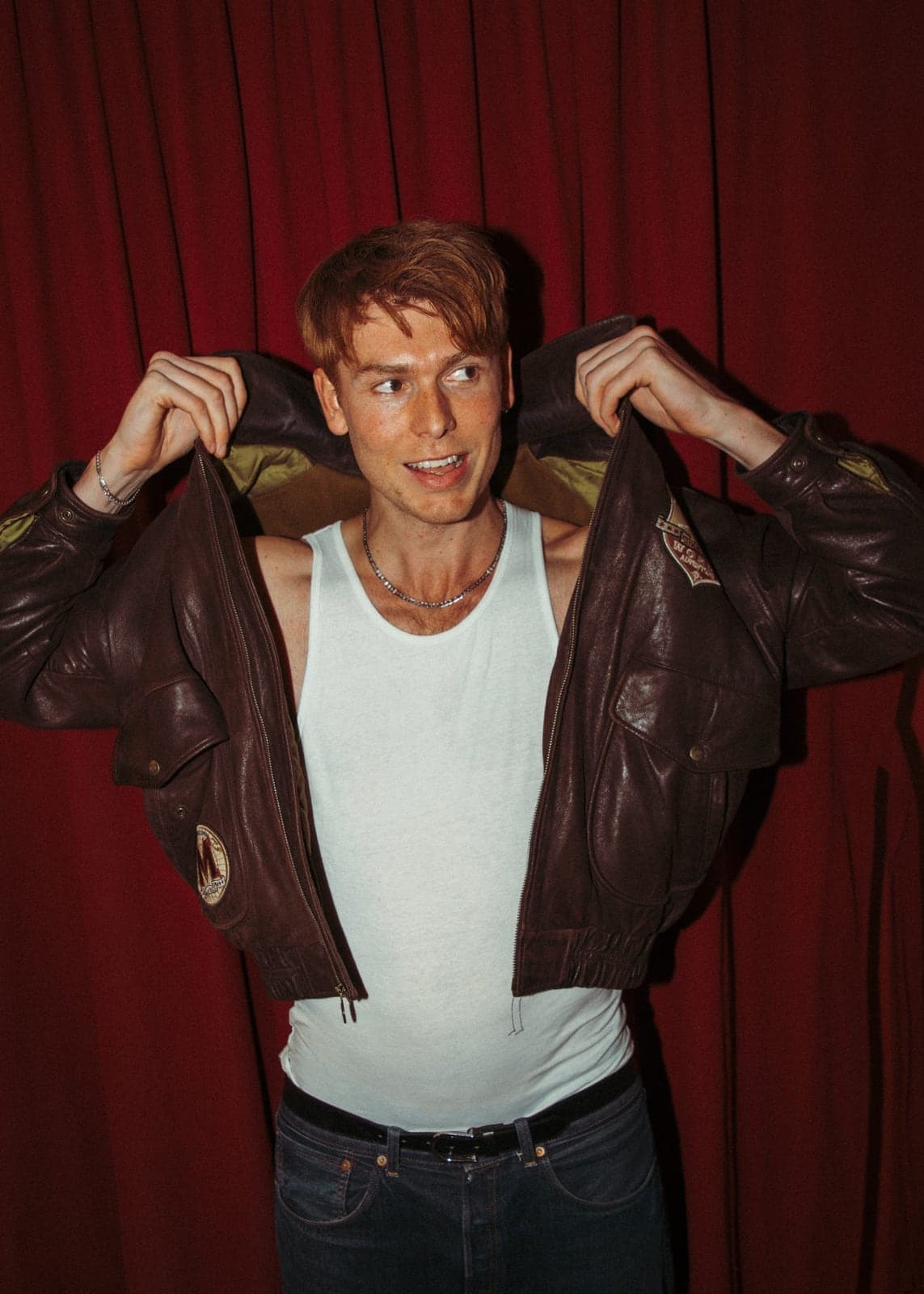 Dan Whitlam holding open a brown leather jacket in front of a red curtain.
