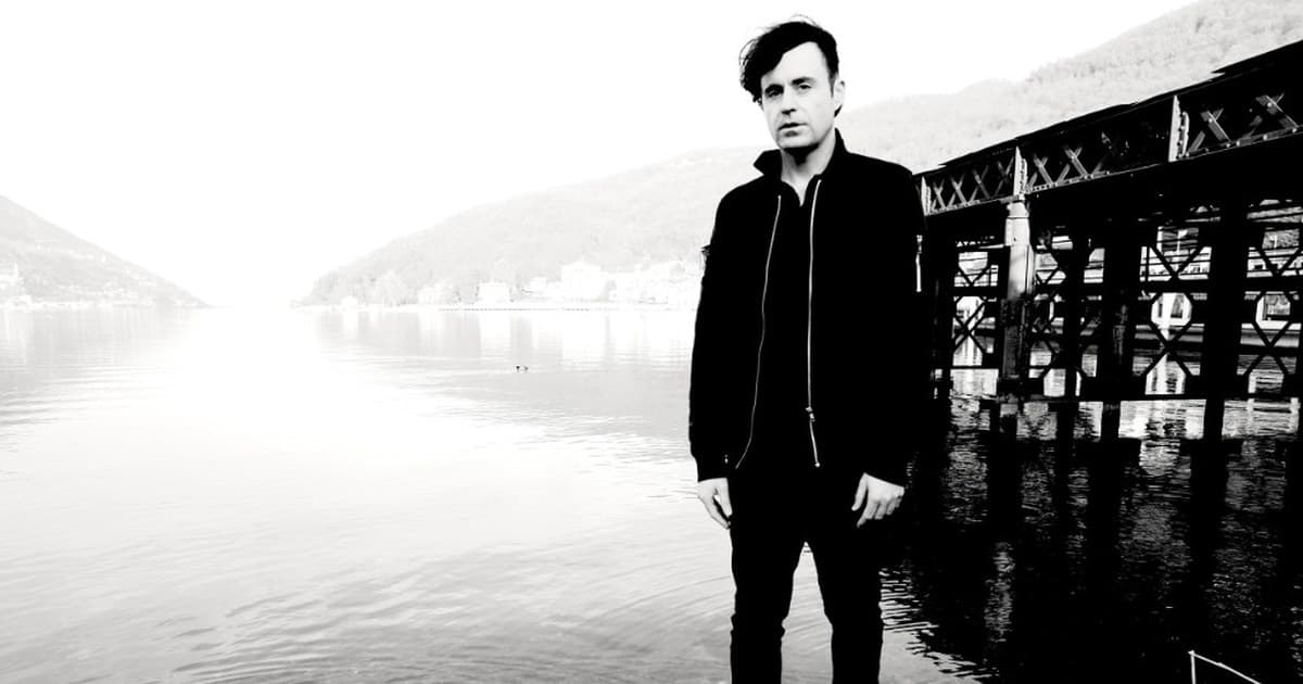 Janosch Moldau standing in shallow water in a black-and-white portrait, with a bridge structure and bright shoreline behind him.