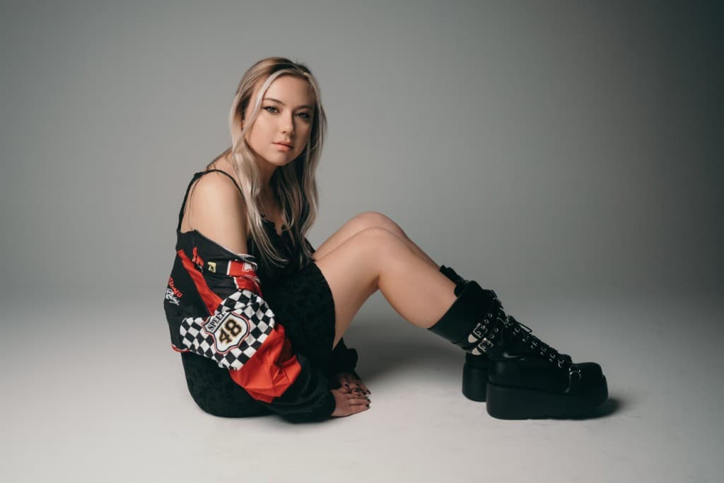 Lauren Presley seated on a studio floor wearing a racing jacket and platform boots.
