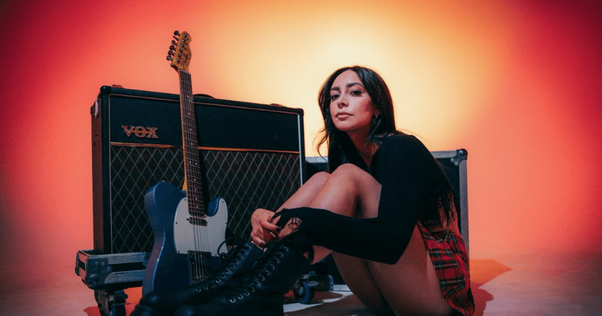 LØLØ seated on a studio floor against a glowing red and orange backdrop, leaning against a Vox amp with a blue Telecaster guitar, wearing a long-sleeved black knit, a red plaid mini skirt, and chunky black combat boots.