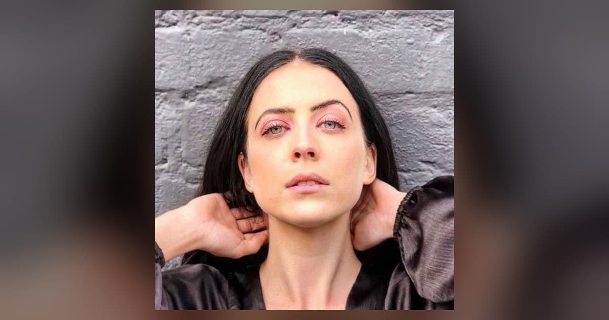 Madalen Duke in a close-up portrait against a gray brick wall, dark hair parted in the middle, hands behind her head, pink eyeshadow catching the light.