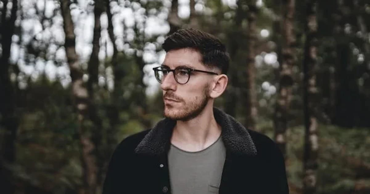 Marsh in a dark jacket and glasses, standing among trees and looking off to the left.