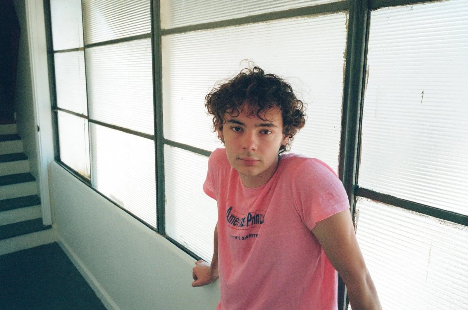 Oscar Lindsey, curly-haired, in a pink T-shirt beside a wall of frosted windows.