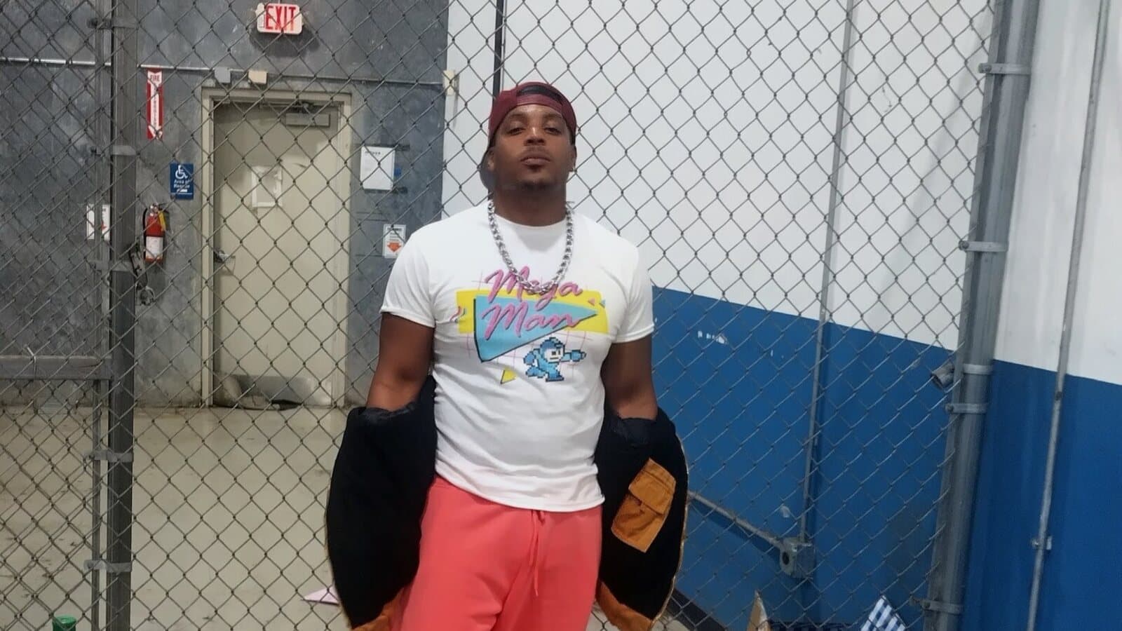 Powers in a white Mega Man T-shirt, coral joggers, a maroon cap, and a silver chain, standing in front of a chain-link fence.