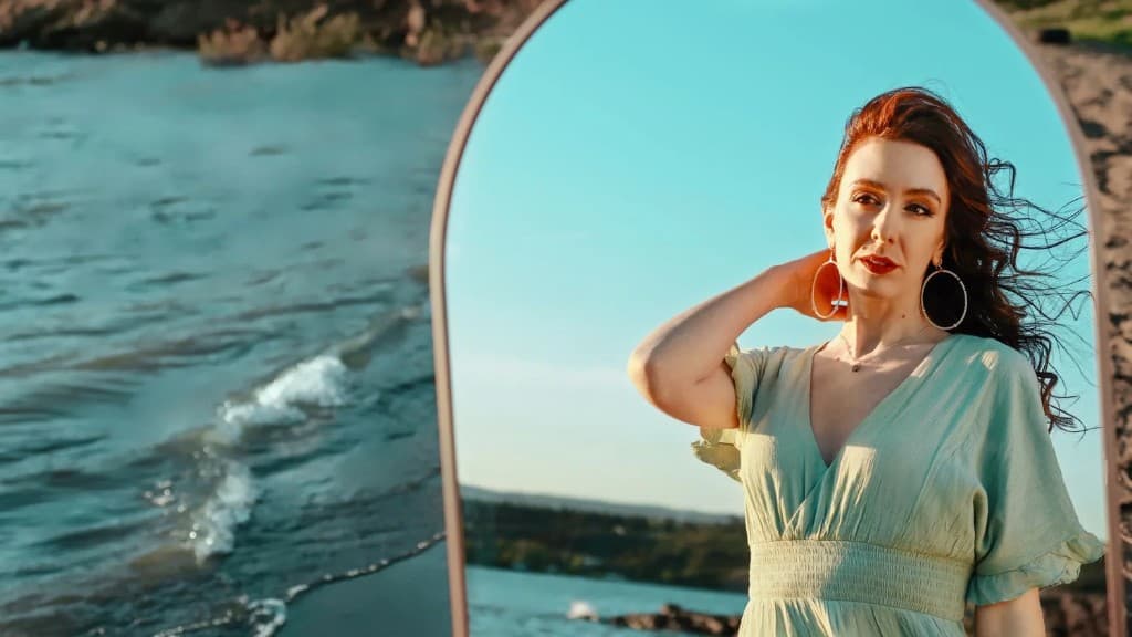 Sabina Chantouria framed inside an arched mirror by the sea, auburn hair lifted by the wind, wearing a sage-green dress and large hoop earrings.