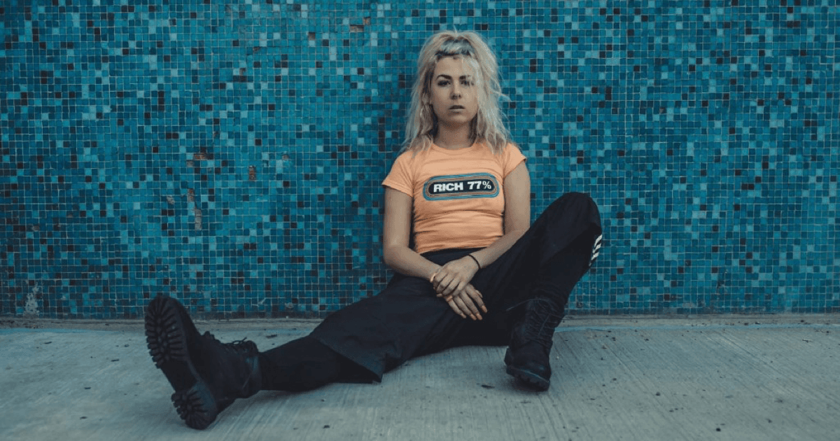 WHIPPED CREAM (Caroline Cecil) seated against a deep blue mosaic-tile wall, blonde hair catching the light, in an orange RICH 77% crop tee, black wide-leg pants, and chunky black boots.