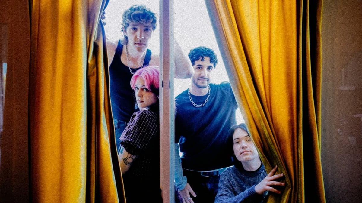 The four members of wilt looking out from between gold curtains in a bright window.