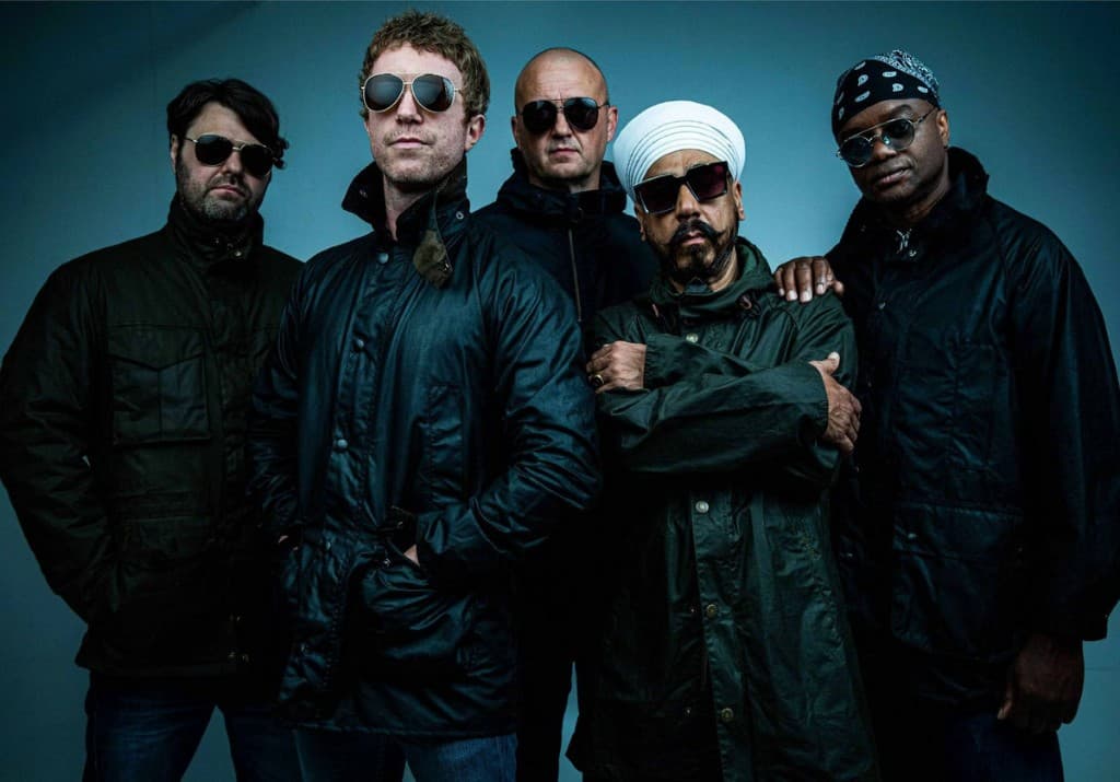 Winachi band portrait. Five members standing together in dark jackets against a moody blue backdrop.